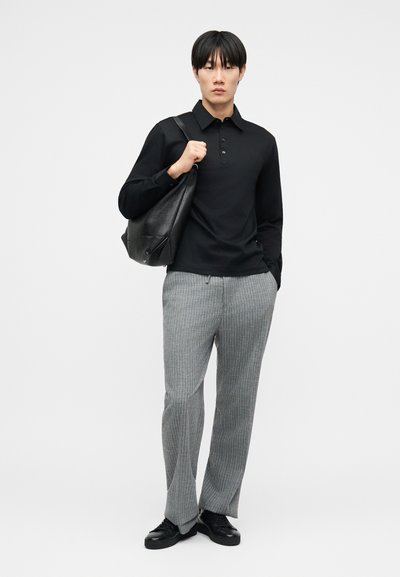 Άνδρας φορώντας μαύρο πικέ πόλο με μακριά μανίκια, γκρι παντελόνι με ρίγες, μαύρα αθλητικά παπούτσια, και κρατώντας μαύρη δερμάτινη τσάντα ώμου, στέκεται μπροστά σε λευκό φόντο.
