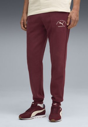 Maroon sweatpants with elastic cuffs, side seams, and a logo print. Worn with matching maroon sneakers featuring a white stripe.