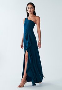 Vestido asimétrico de color verde azulado con volantes en un lado y abertura en el muslo. Hecho de tela suave. Combinado con tacones altos metálicos con tiras.