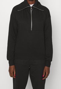 Sweat-shirt noir zippé avec un col montant, une fermeture éclair métallique et des poignets côtelés. Tissu doux avec une coupe décontractée et un design minimal.