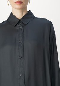 Femme portant une chemise noire ample et brillante avec un col et des boucles d'oreilles pendantes dorées, vue des épaules au menton.