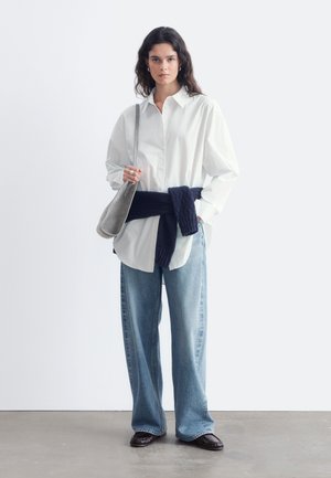 Junge Frau trägt ein lockeres weißes Hemd, weite blaue Jeans, dunklen Pullover um die Taille gebunden, schwarze Schuhe und eine graue Schultertasche und steht vor einer schlichten Wand.
