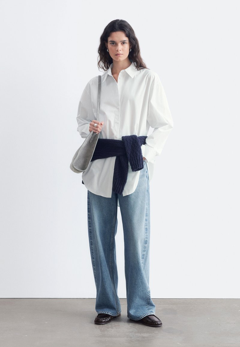 Jeune femme portant une chemise blanche ample, un jean bleu à larges jambes, un pull sombre noué autour de la taille, des chaussures noires et un sac à épaule gris, debout contre un mur uni.