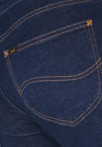 Un bolsillo de jeans de mezclilla azul oscuro con costuras amarillas visibles y una etiqueta negra con "Lee." Textura suave y diseño curvado.