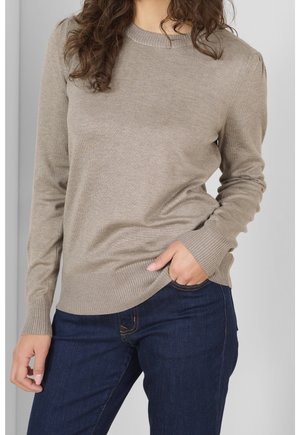 Femme portant un pull beige à manches longues et un jean bleu foncé, avec une main dans la poche et les cheveux bouclés partiellement visibles.