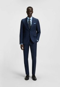Completo blu navy su misura con rever a pizzetto, due bottoni e pantaloni su misura. Camicia azzurro chiaro, cravatta a righe e dettaglio di fazzoletto da taschino.