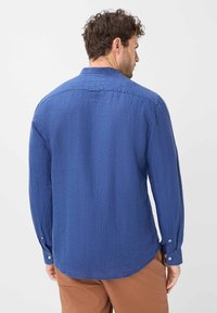 Camicia blu a maniche lunghe con un tessuto texturizzato, orlo curvo e dettagli con bottoni sulle maniche, abbinata a pantaloni marroni.