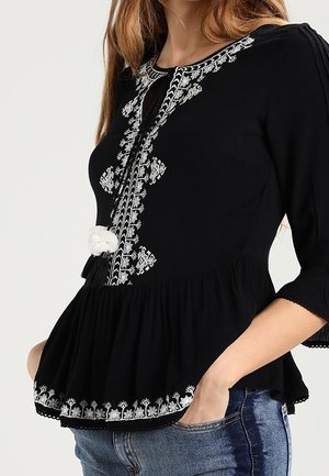Femme portant un top péplum noir avec broderie florale blanche et pompons, associé à un jean bleu, une main dans la poche.