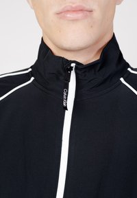 Giacca nera con colletto alto, caratterizzata da strisce bianche decorative e chiusura a zip. Etichetta con il logo visibile vicino al collo. Texture liscia.