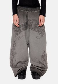 Άτομο που φοράει φαρδιά γκρι παντελόνια με μαύρα διακοσμητικά σχέδια και μαύρα αθλητικά παπούτσια, μπροστά σε απλό φόντο.