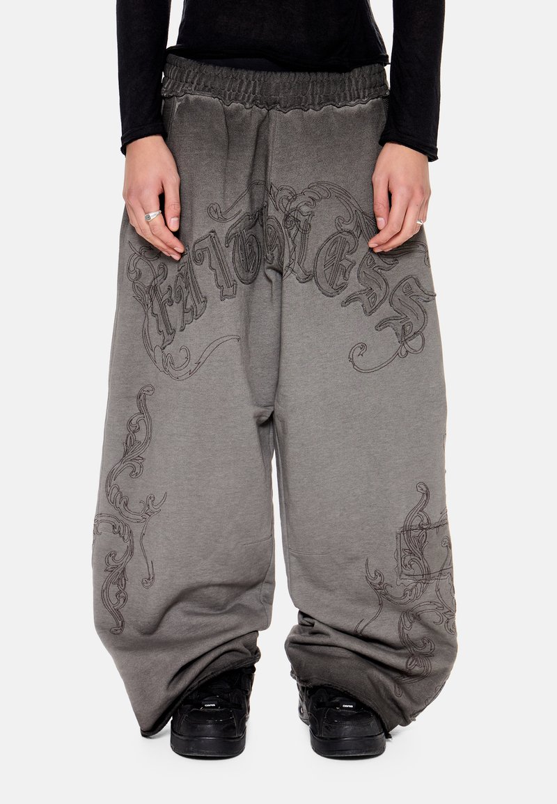 Άτομο που φοράει φαρδιά γκρι παντελόνια με μαύρα διακοσμητικά σχέδια και μαύρα αθλητικά παπούτσια, μπροστά σε απλό φόντο.