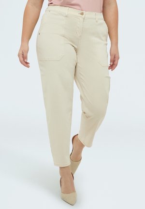 Kvinde iført beige bukser med høj talje og forlommer samt beige spidse hæle, gående mod en ensartet lys baggrund.