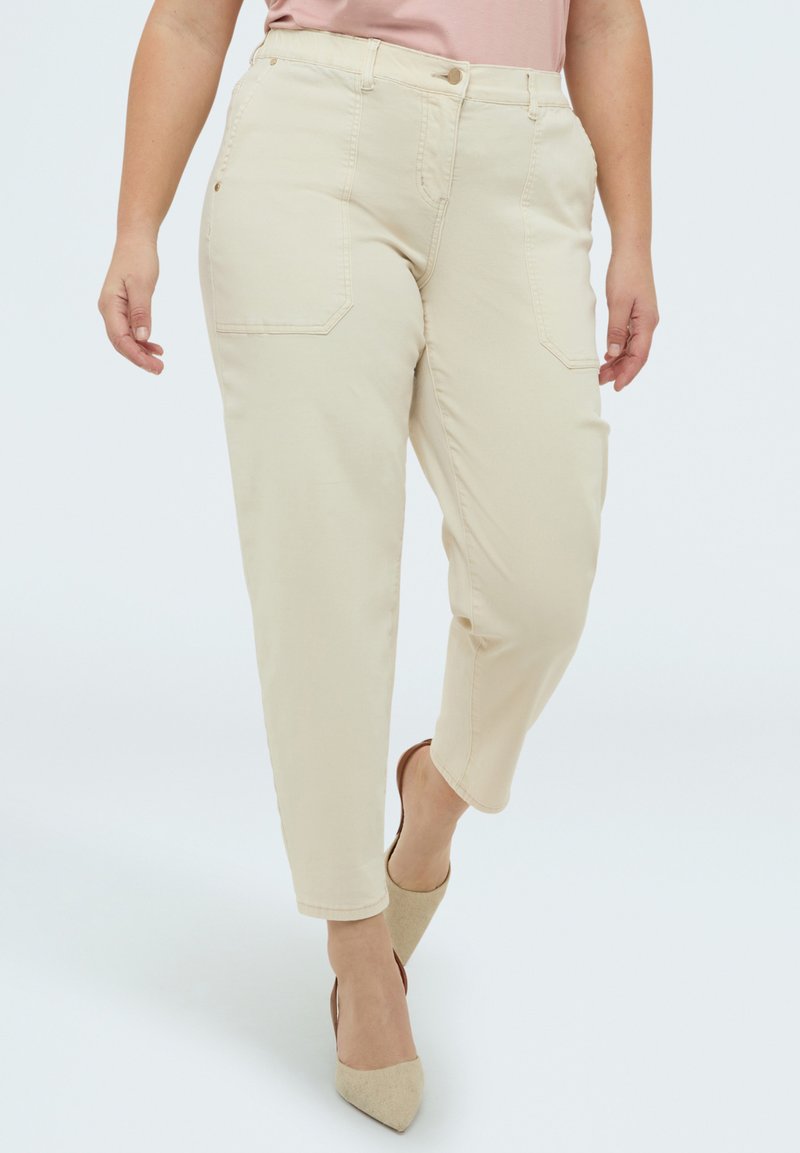 Femeie purtând pantaloni bej cu talie înaltă, cu buzunare frontale, și tocuri bej cu vârf ascuțit, mergând pe un fundal simplu, deschis la culoare.