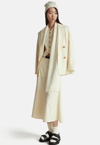 Krémový oblek s dvoudílným sakem, odpovídající košile, dlouhá sukně a pletený šátek. Doplňují ho černé sandály a bílé ponožky.