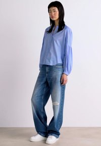 Chemise bleu clair à manches bouffantes, assortie à un jean bleu délavé coupe ample et des baskets blanches, portée par un mannequin sur fond neutre.