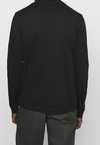 Bakifrån av en person som bär en svart långärmad topp och mörkgröna byxor, stående mot en enkel ljus bakgrund.