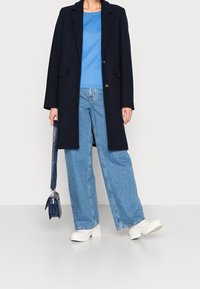 Cappotto in misto lana navy, maglione azzurro chiaro, jeans larghi in denim chiaro, stivali bianchi chunky e una piccola borsa navy con tracolla.
