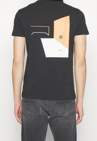 Camiseta de algodón negra con un diseño abstracto en blanco y naranja en la parte de atrás, con formas geométricas y un acento circular.