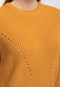 Käytännöllinen neulepaita sinapinkeltaisena, jossa on pyöreä kaula-aukko, teksturoitu kuvio ja vinot aukot vasemmalla puolella.