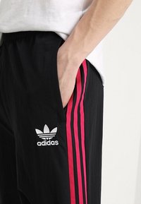 Černé sportovní kalhoty se třemi růžovými pruhy na bocích a bílým logem Adidas umístěným blízko kapsy.