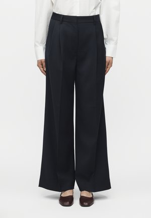 Γυναίκα που φοράει μαύρο παντελόνι με φαρδιά πόδια, λευκό πουκάμισο με μακριά μανίκια και σκούρα καφέ παπούτσια με τετράγωνη μύτη, στέκεται μπροστά σε ένα απλό φόντο.