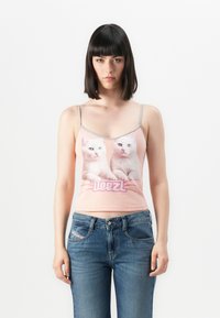 Ung kvinne med strakt mørkt hår iført en rosa cami med hvite kattetrykk og blå jeans, stående mot en ensfarget hvit bakgrunn.