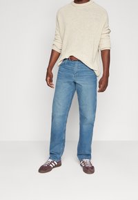 Helles beiges Strickpullover mit hochgeschlagenen Ärmeln, kombiniert mit hellblauen geraden Jeans und dunkelvioletten Sneakers mit weißen Streifen.