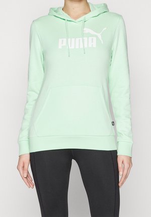 Osoba stojící na jednobarevném pozadí, oblečená ve světle zelené mikině Puma s přední kapsou a černých legínách.