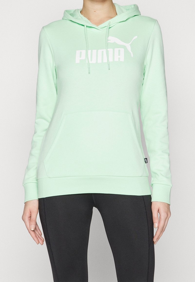 Personne portant un sweat à capuche Puma vert clair avec poche avant et des leggings noirs, debout devant un fond uni.