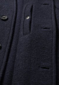 Close-up van de donkere wollen stof van een jas met zwarte knopen, knoopsgaten en een klepzak die is vastgezet met een metalen drukknoop.