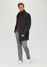 Donkergrijze jas van gestructureerde stof, met hoge kraag en een ritszak op de borst, gedragen over een kastanjebruine shirt en grijze tapered broek met witte sneakers.