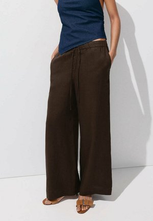 Persona con pantalones anchos marrón oscuro, top sin mangas azul marino y sandalias abiertas color beige, de pie con las manos en los bolsillos.