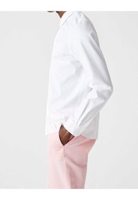 Camisa blanca de botones con mangas largas, cuello puntiagudo y botones en los puños, combinada con pantalones rosa claro. Textura suave, corte ajustado.