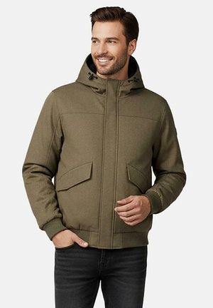Man die lacht en een olijfgroene capuchonjas met voorzakken en een zwarte broek draagt tegen een lichte, effen achtergrond.
