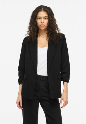 Göndör hajú nő fekete blézert visel fehér felső és fekete nadrág felett, egyszerű fehér háttér előtt áll.