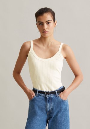 Junge Frau mit dunklen, zurückgebundenen Haaren, trägt ein cremefarbenes, geripptes Tanktop, blaue Jeans und einen schwarzen Gürtel, Hände in den Taschen, neutraler Hintergrund.