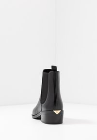 Stivaletto Chelsea in pelle nera con pannelli laterali elastici, punta affusolata e tacco basso, caratterizzato da un accento triangolare dorato sul retro.