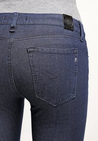 Vaqueros de mezclilla azul oscuro con un bolsillo trasero que presenta un diseño de chevrón bordado y una etiqueta metálica de la marca en la cinturilla.