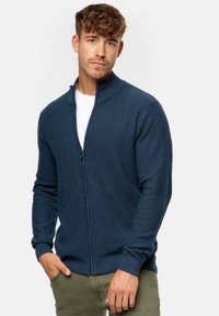 Cárdigan azul marino con cremallera en tejido de punto texturizado, con cuello alto y puños acanalados, llevado sobre una camiseta blanca.