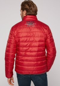 Giacca rossa gonfiabile con colletto a coste, presenta sezioni trapuntate orizzontali e dettagli sul retro con la scritta: "2501 ALASKA WATER & ICE CAMP DAVID."