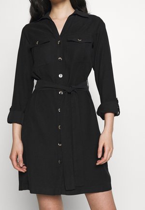 Kobieta w czarnej sukience zapinanej na guziki z kieszeniami na piersi, z podwiniętymi rękawami i wiązanym materiałowym paskiem w talii.