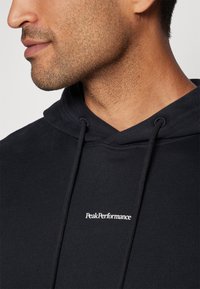 Svart hettegenser laget av mykt materiale med hette og hvit "PeakPerformance"-logo på brystet. Synlig teksturert stoff og sømdetaljer.
