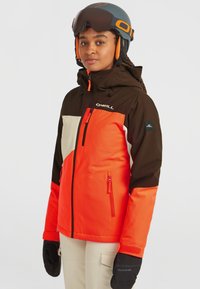 Personne portant une veste de ski bicolore marron, orange et beige, des gants noirs et un casque de ski noir avec des accents oranges.