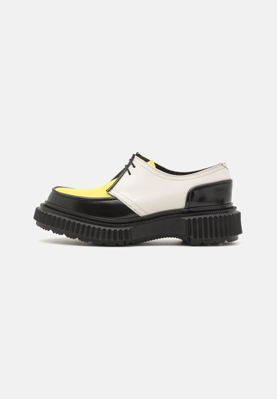 Schwarz-weiße Schuhe mit einer dicken Sohle; verfügen über einen gelben Akzent an der Spitze und strukturierte schwarze Gummisohlen für besseren Halt.