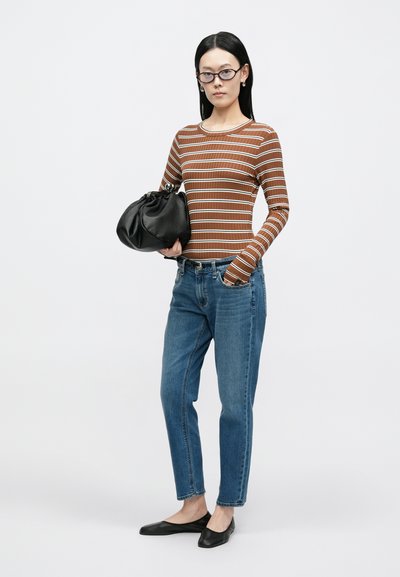 Pruunide triipudega pika varrukaga top, sinised teksapüksid, mustad madalad kingad ja must volditud käekott. Modell seisab valge tausta ees.