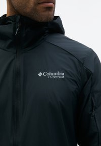 Muž v čiernej bunde Columbia Titanium s predným zipsom a vreckom na zips na rukáve, orezaný pri brade a ramene.