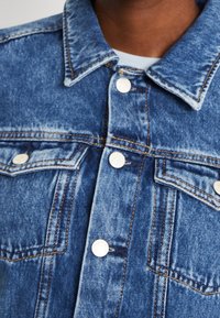 Denim jacket featuring a blue wash, light distressing, silver snap buttons, and two chest pockets with contrasting stitching details.
