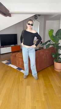 Musta avonainen pitkähiha, vaaleansiniset levennetyt farkut, beiget kengät, sisätila, taustalla kasveja ja huonekaluja.