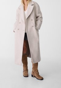 Donna che indossa un lungo cappotto beige doppiopetto sopra un maglione bianco e una gonna nera, abbinato a stivali in suede marrone al ginocchio.