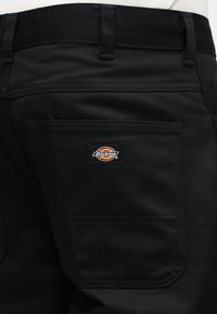 Gros plan sur la poche d'un pantalon noir avec un patch logo de la marque Dickies centré sur la poche.